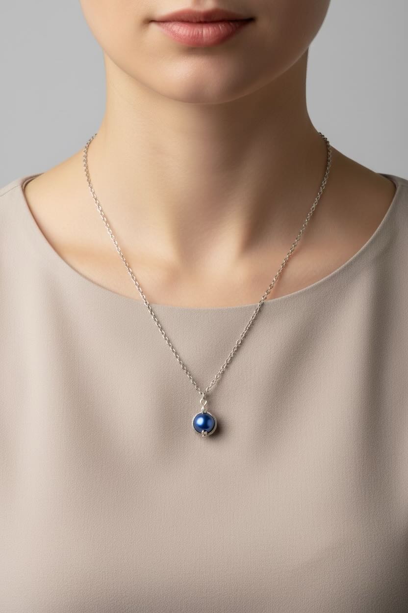 A close-up of a model wearing a delicate silver chain necklace with a small, round, dark royal blue pearl pendant.