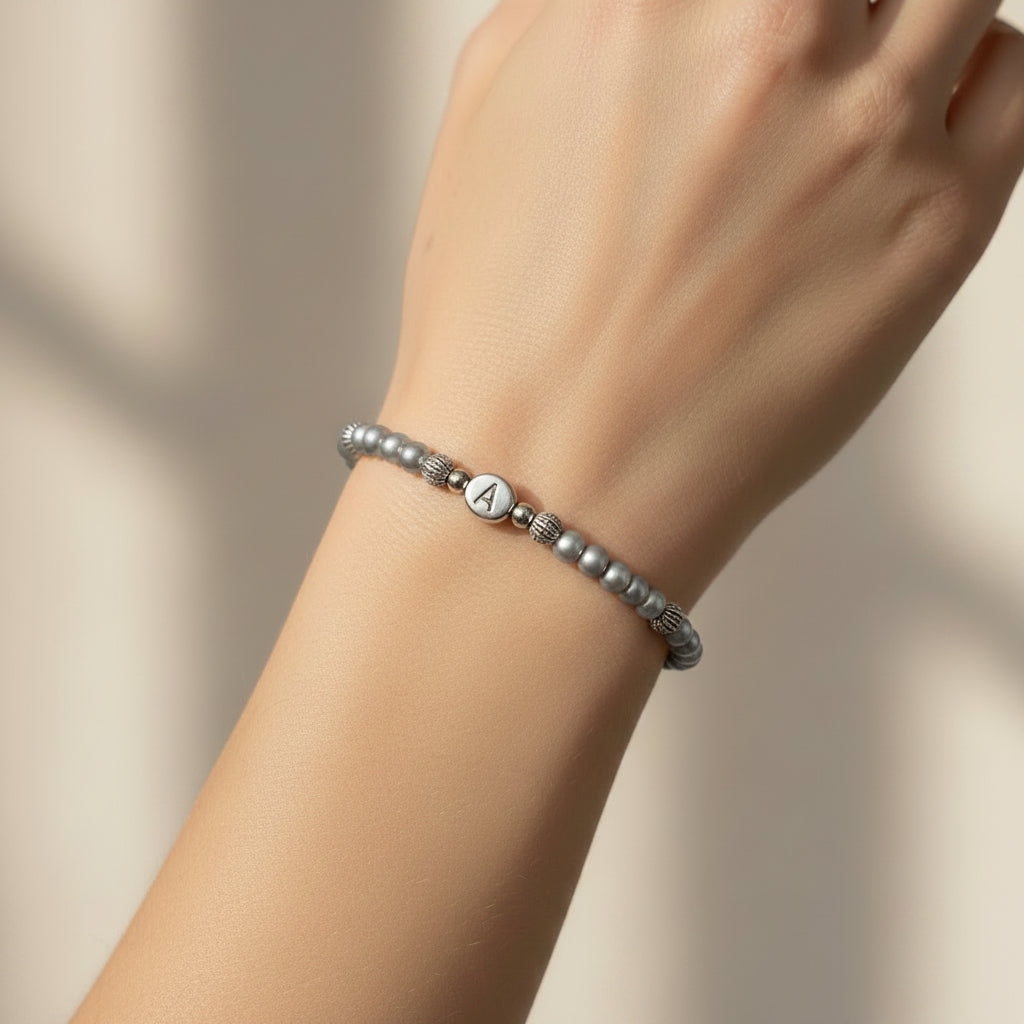 A silver grey pearl bracelet displayed on a model's arm. The bracelet features silver grey glass pearls, small silver spacer beads, and a central round silver bead engraved with letter A.