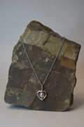 A silver heart pendant necklace with a lavender pearl center, draped over a textured  stone block against a neutral gray background.