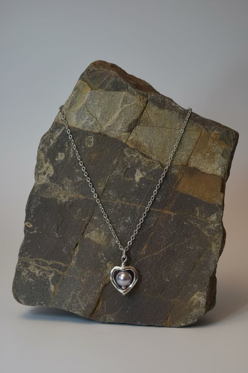 A silver heart pendant necklace with a lavender pearl center, draped over a textured  stone block against a neutral gray background.