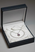 A silver chain necklace with a round lavender pearl pendant displayed inside an open black presentation box lined with white tissue paper.