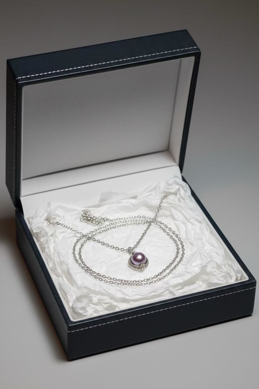 A silver chain necklace with a round lavender pearl pendant displayed inside an open black presentation box lined with white tissue paper.