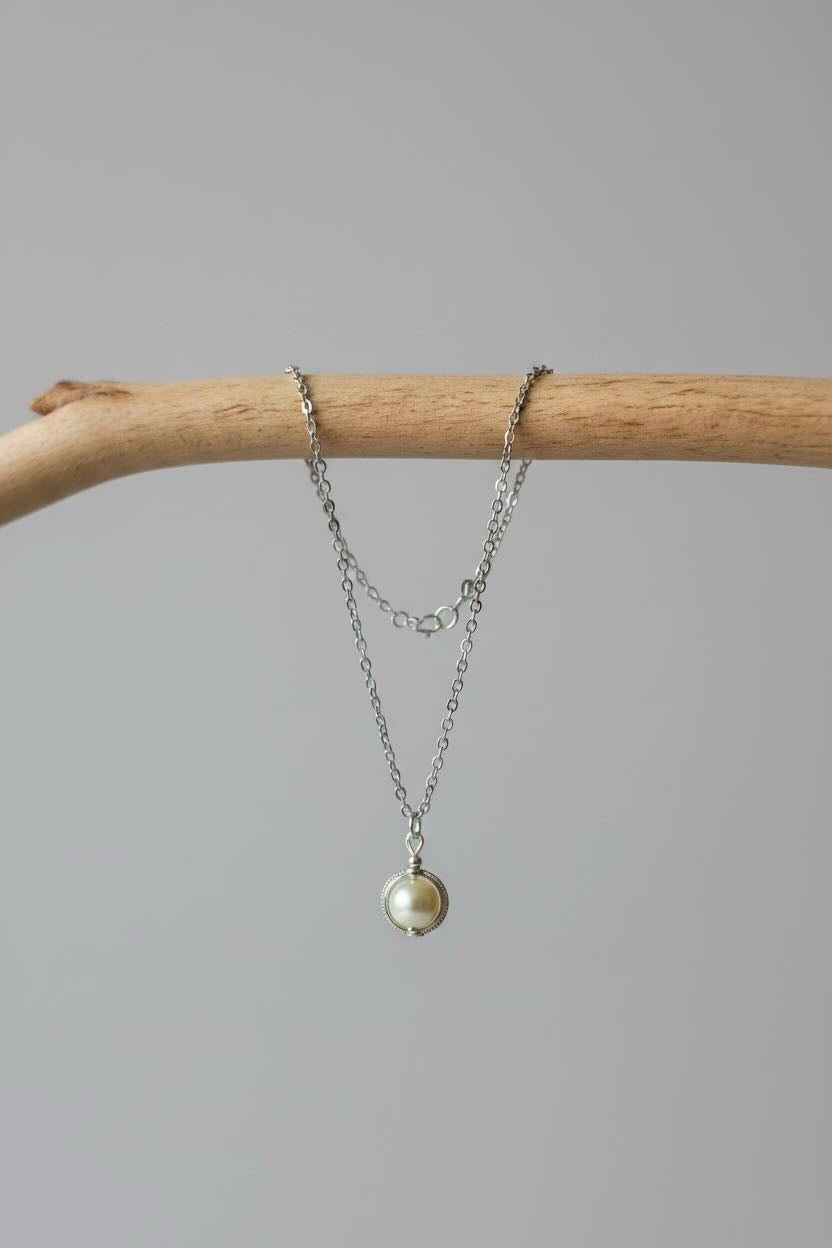 A delicate silver necklace with a small round pale cream pearl pendant, draped over a light brown wooden branch against a solid gray background.