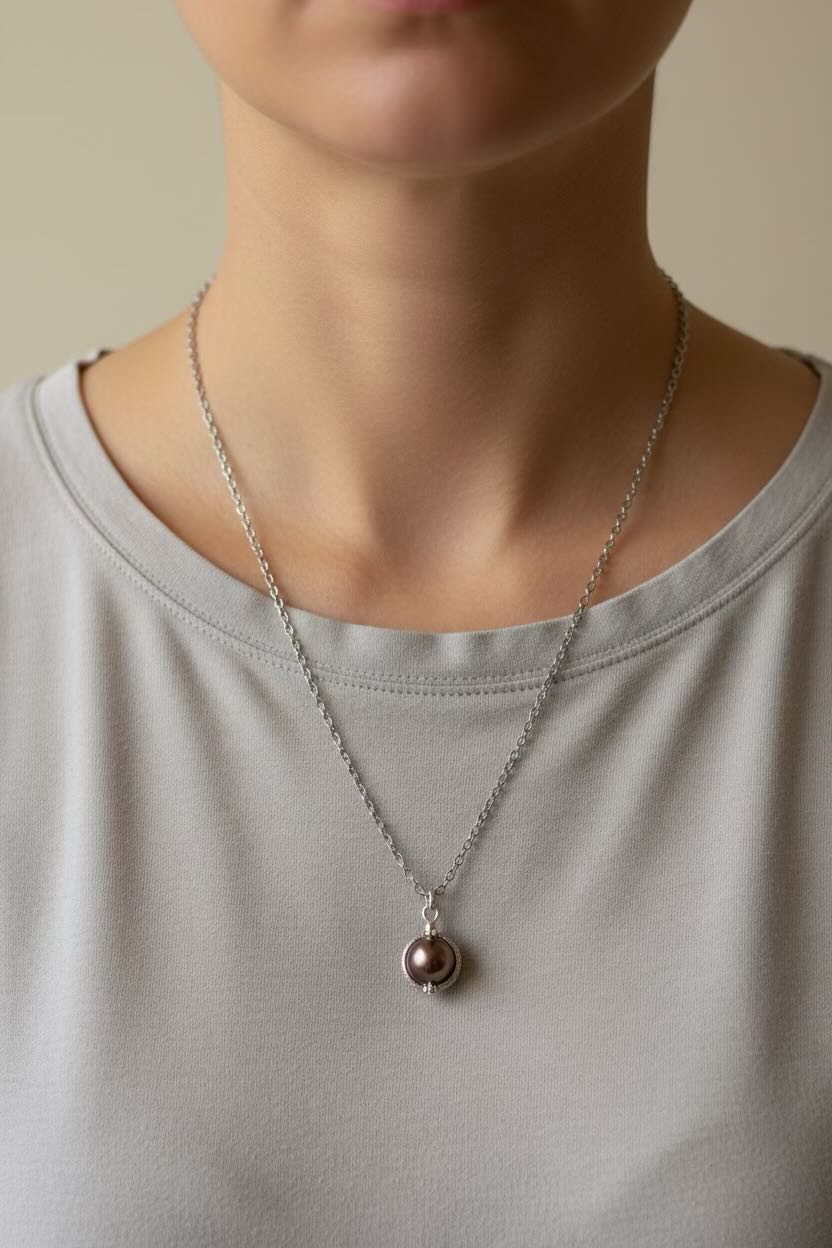 A close-up of a person wearing a delicate silver chain necklace with a small round pendant featuring a saddle brown pearl, paired with a light gray crew-neck top.