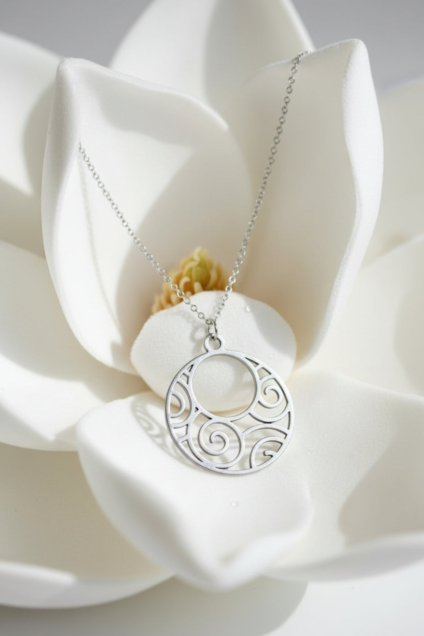 A silver pendant necklace with a circular cutout design featuring swirl patterns, displayed on a white petal flower.