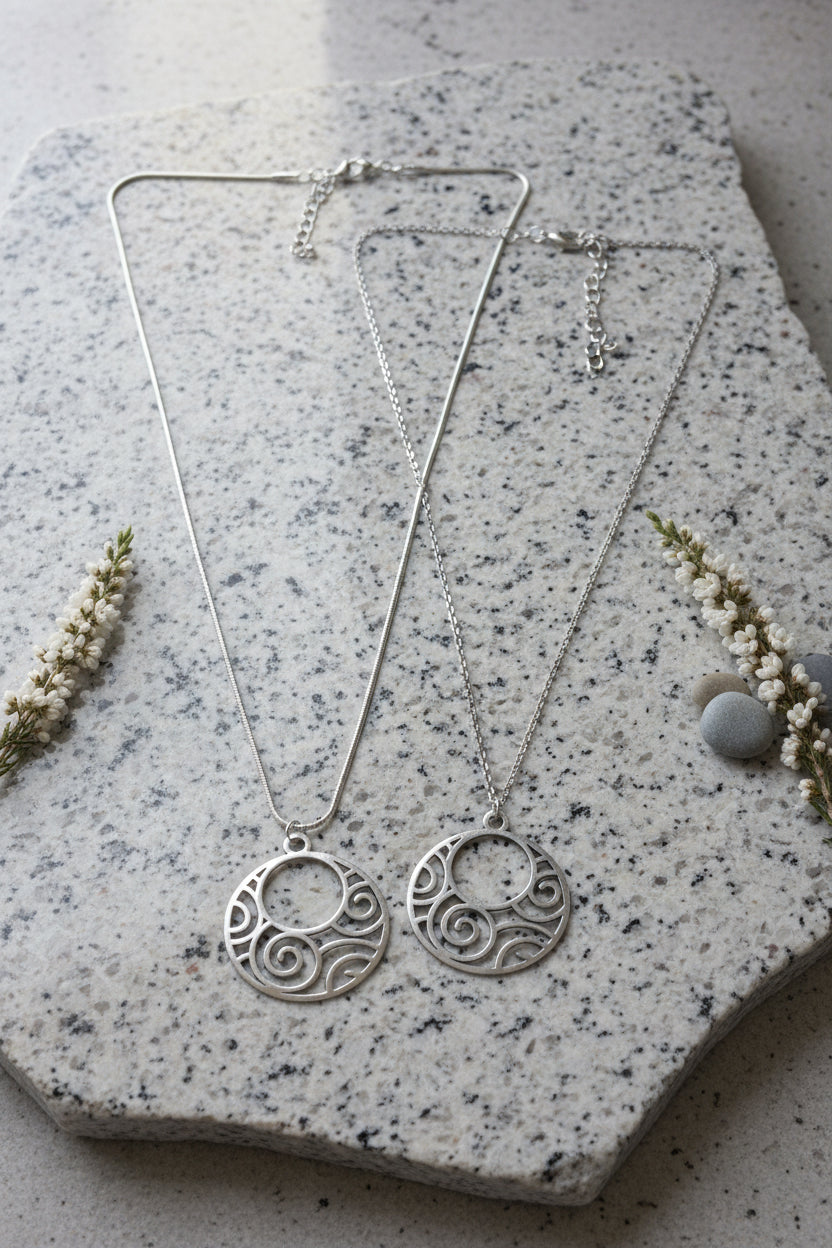 Two silver-tone circular pendant necklaces with openwork swirl patterns, displayed on a large silver grey stone.