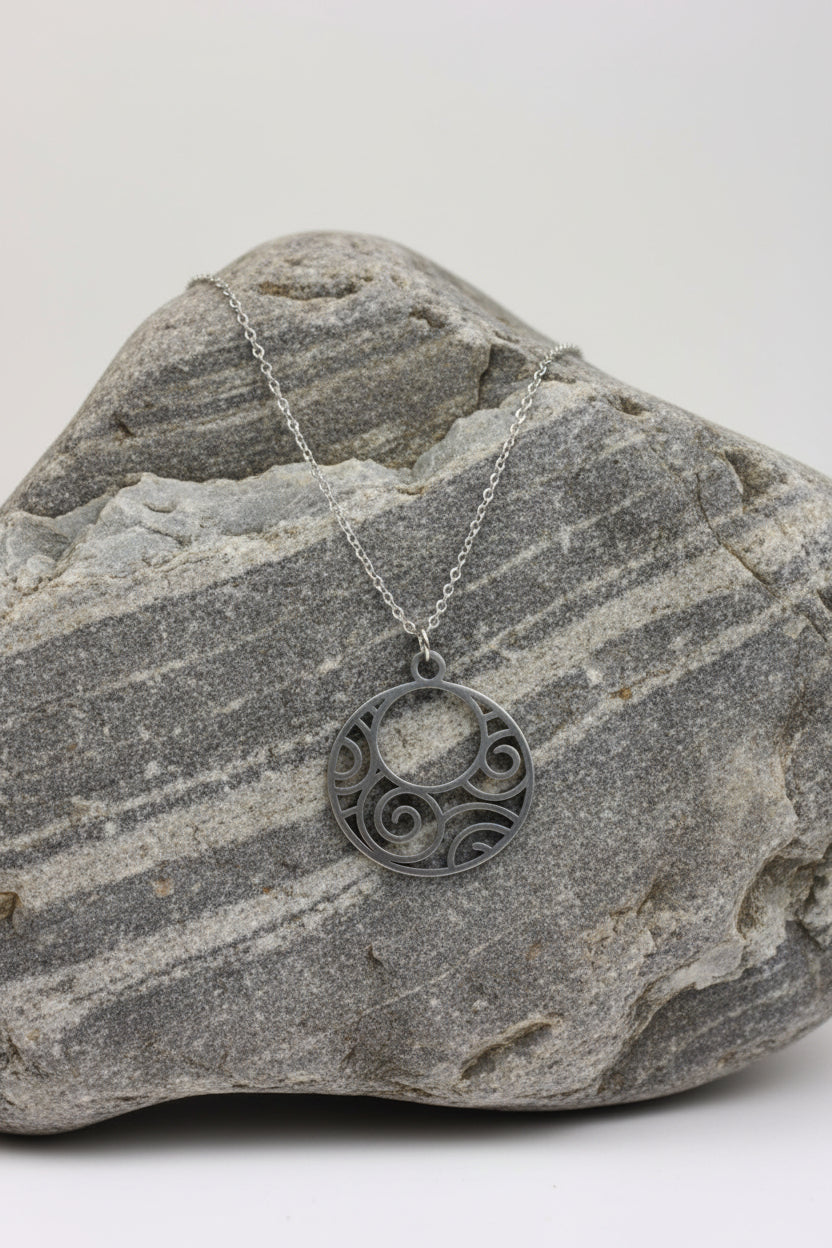 A silver pendant necklace with a circular cutout design featuring swirl patterns, displayed on a grey stone.