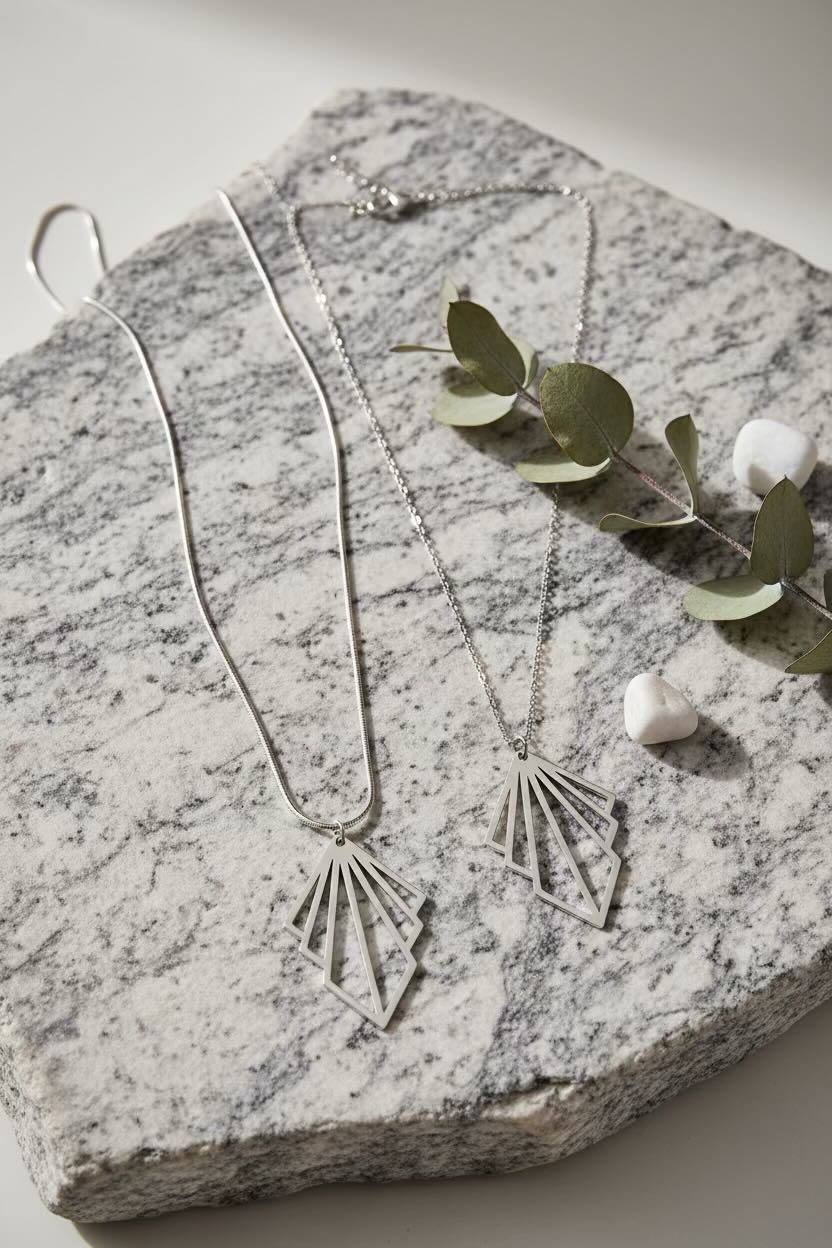Two silver art deco fan pendant necklaces displayed on a textured gray stone slab with eucalyptus leaves and small white stones.