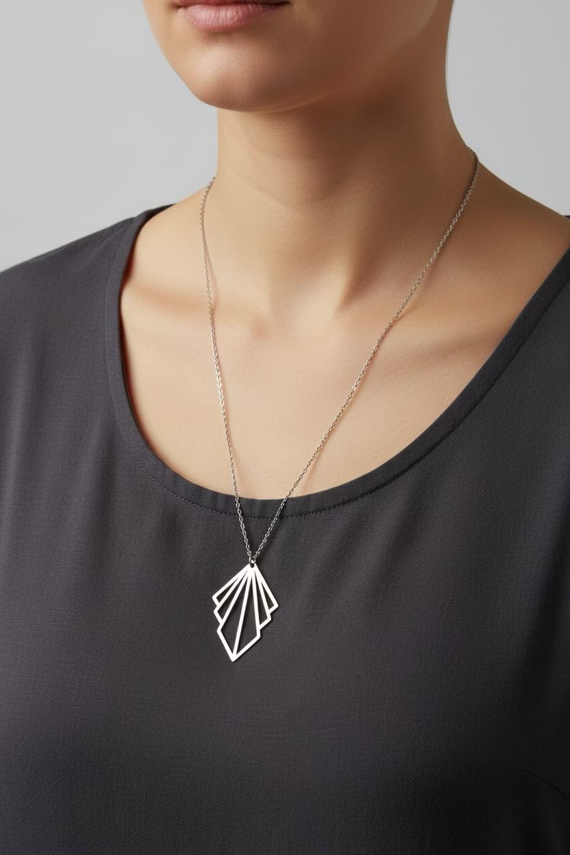 A close-up of a model wearing a delicate silver necklace with a geometric fan-shaped pendant, paired with a dark gray scoop-neck top against a neutral gray background.