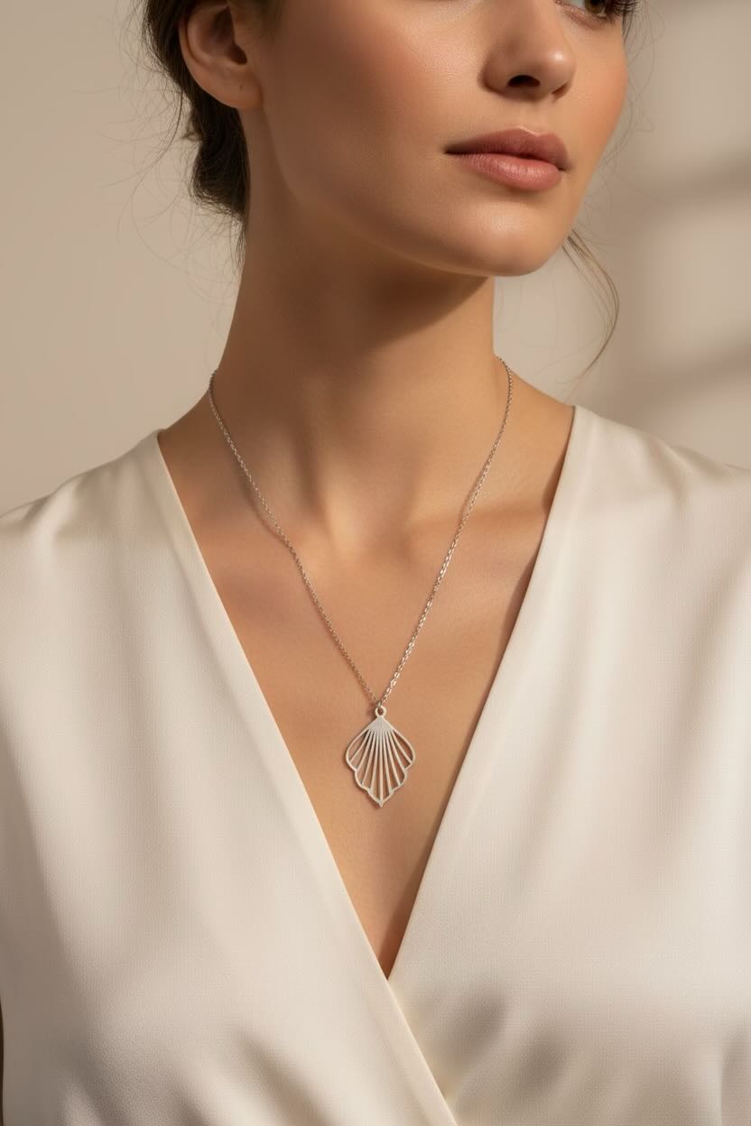 A close-up of a woman wearing a delicate silver chain necklace with a fan-shaped pendant, styled with a cream satin V-neck top against a warm beige background.