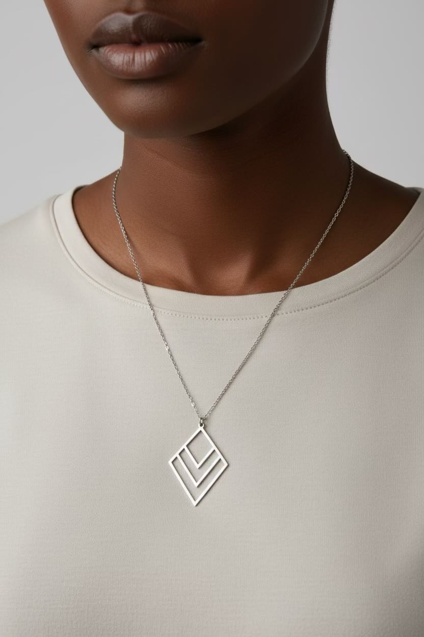 A close-up of a model wearing a silver layered diamond chevron pendant necklace with a diamond-shaped interlocking design, paired with a cream-colored top against a neutral gray background.