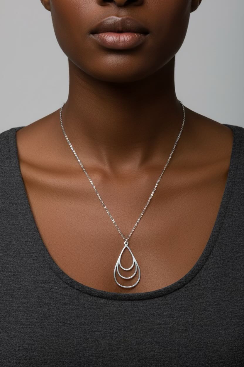 A close-up of a model wearing a silver necklace with a teardrop-shaped pendant made of three interlocking open loops, paired with a dark gray scoop-neck top against a neutral gray background.