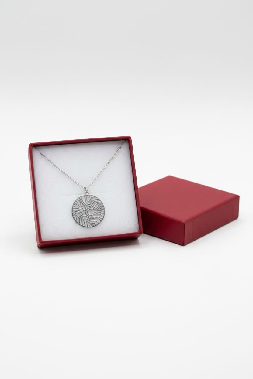 A silver pendant necklace with a textured, circular wave pattern circle pattern, presented in an open red gift box with a white insert next to its closed red lid on a light gray background.