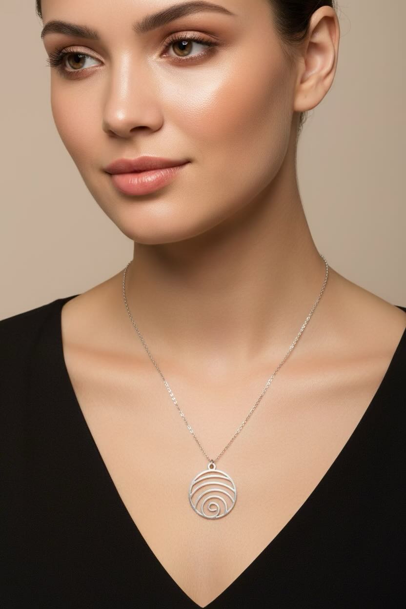 A close-up of a woman wearing a delicate silver necklace with a circular spiral shell pendant, styled with a black V-neck top against a soft beige background.