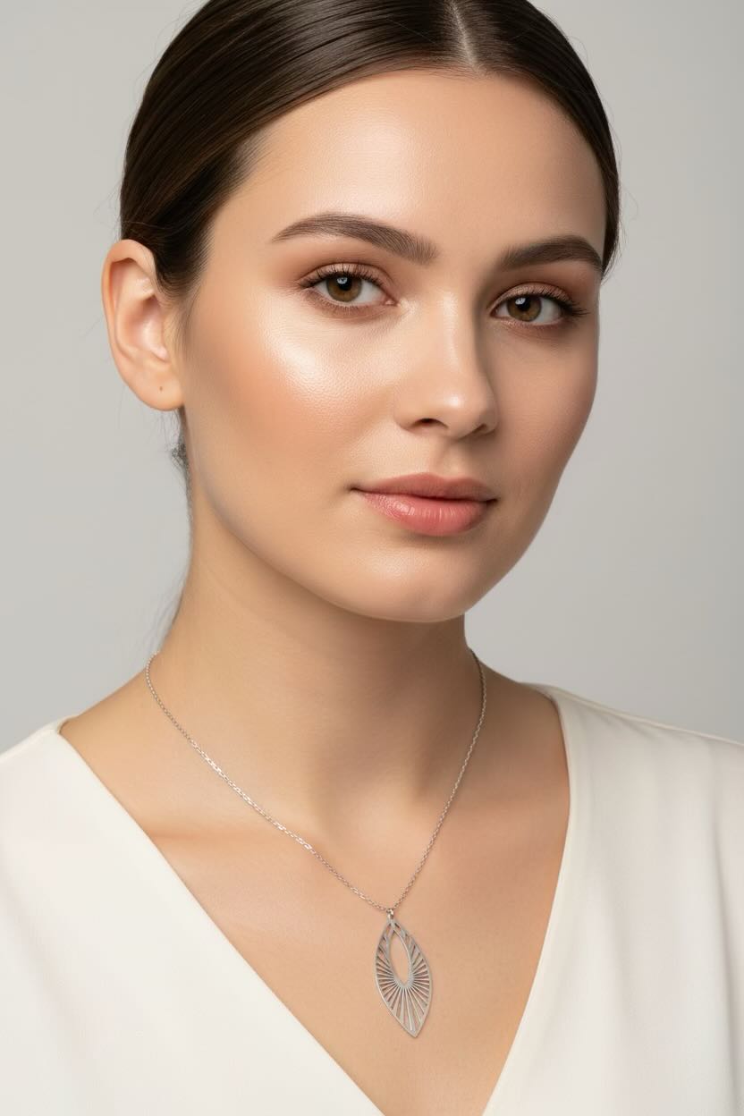 A close-up portrait of a woman wearing a delicate silver chain necklace with a sunburst marquise-shaped pendant featuring a sunburst or fan pattern, set against a clean, light gray background.