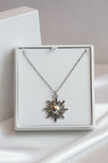 A silver sunburst pendant necklace with a central pearl, presented in a white square gift box on a soft white background.