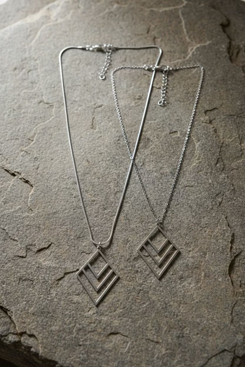 A pair of silver layered diamond chevron pendant necklaces with interlocking diamond and chevron designs, displayed on a textured gray stone surface.