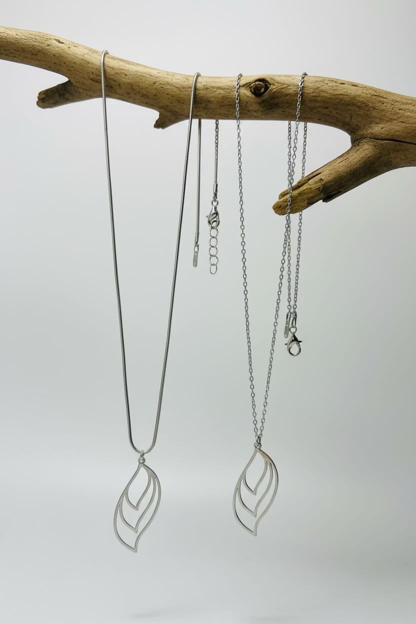 Three silver-toned leaf-shaped pendant necklaces hanging from a natural wood branch against a light gray background.