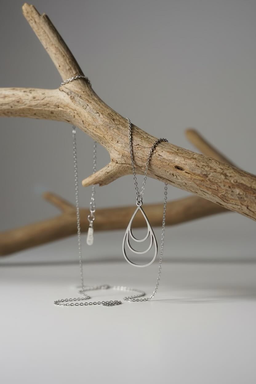A silver-toned layered teardrop pendant necklace draped over a natural wood branch against a neutral gray background.