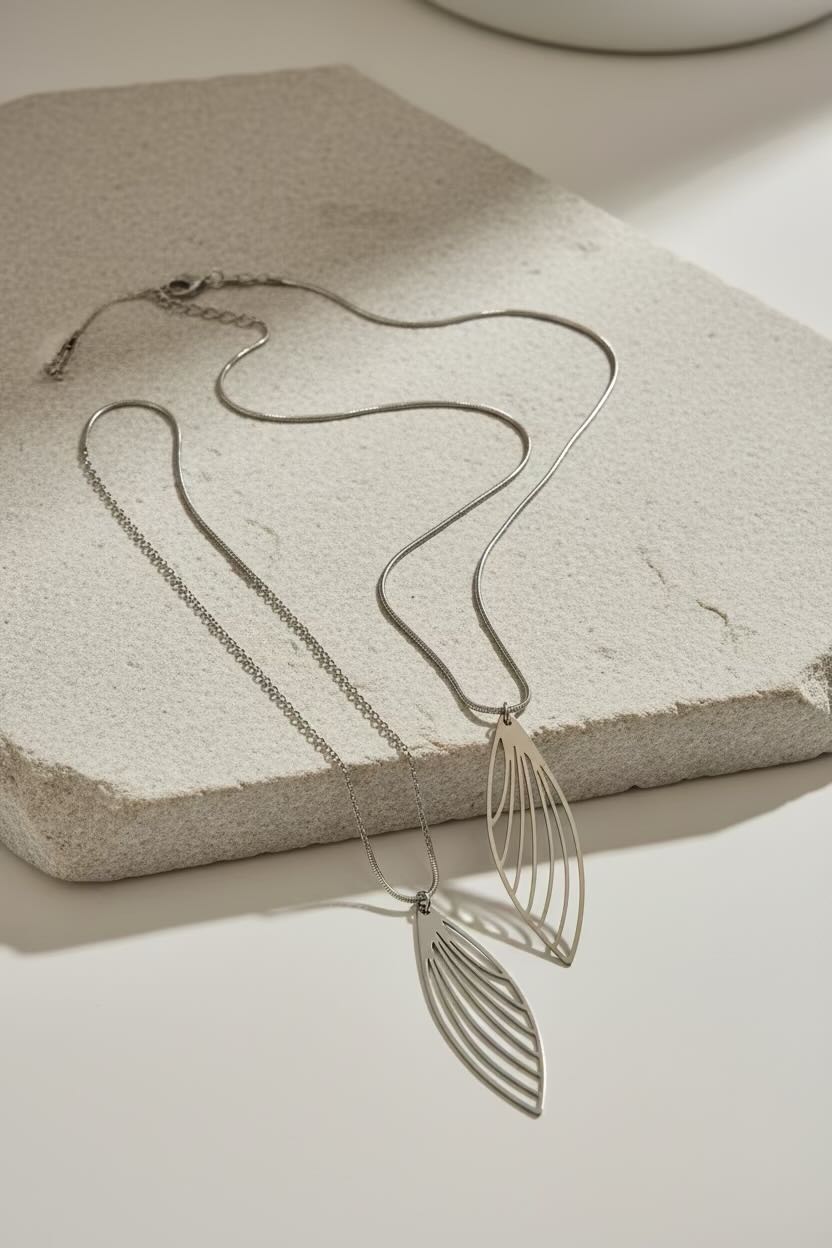 A silver-toned necklace with a delicate chain and a large openwork feather wing-shaped pendant, displayed on a textured light stone slab against a warm off-white background.