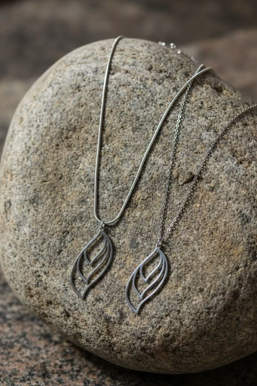 Two silver leaf-shaped pendant necklaces draped over a large smooth stone, with one pendant fully visible and the other partially folded.