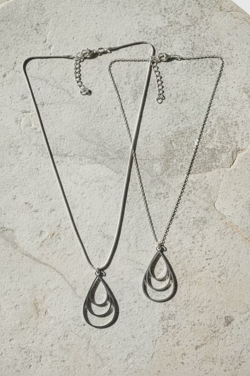Two silver-toned teardrop pendant necklaces with open-link designs, displayed on a textured light beige stone surface.