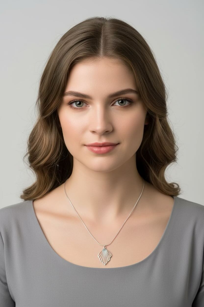 A woman with brown hair wearing a silver necklace with a fan shaped pendant, photographed against a light gray background.