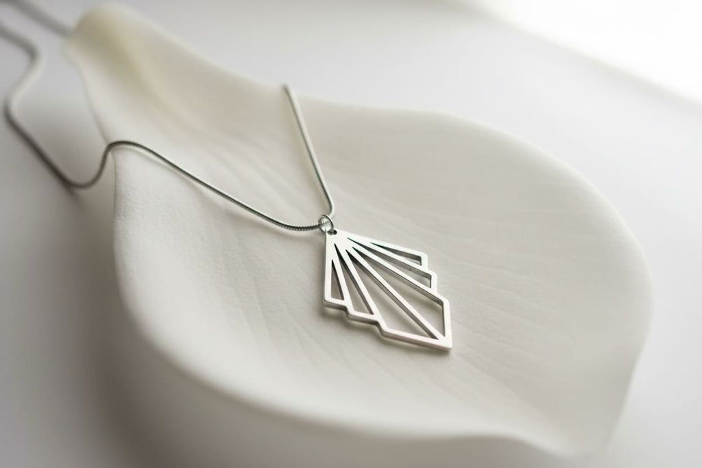 A silver art deco fan pendant necklace with a fan-like openwork design, resting on a white ceramic dish against a soft white background.
