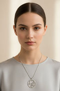 A close-up portrait of a woman wearing a delicate silver pendant necklace with a circular geometric design, paired with a light gray top against a soft beige background.