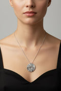 A close-up of a model wearing a silver pendant necklace with a textured circular design, paired with a black top against a neutral gray background.
