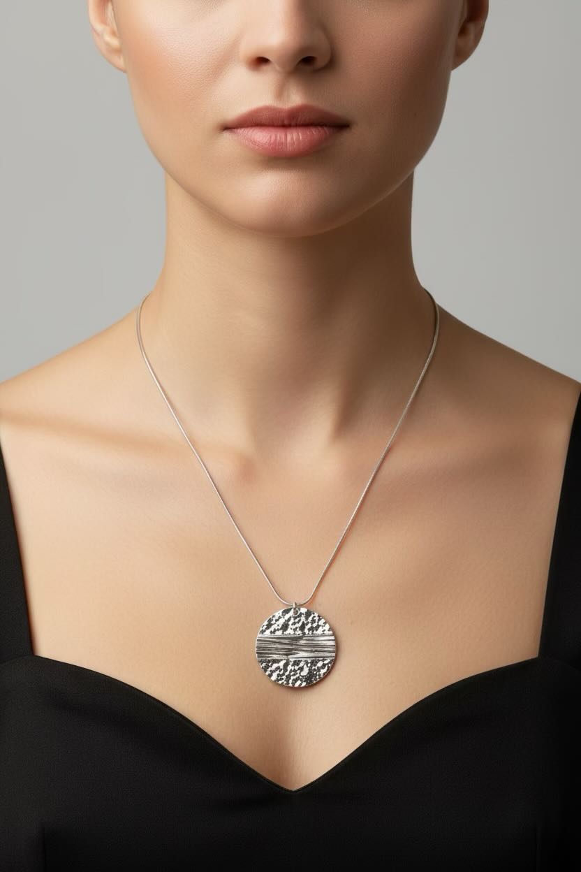 A close-up of a model wearing a silver pendant necklace with a textured circular design, paired with a black top against a neutral gray background.