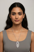 A woman with dark hair wearing a silver pendant necklace with a sunburst design, paired with a gray sleeveless top, against a neutral gray background.