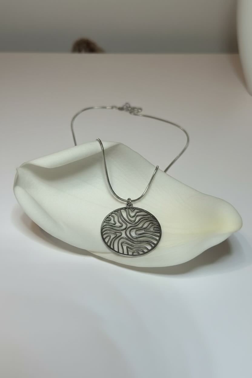 A silver necklace with a circular pendant featuring a textured, wavy damascus-style pattern, displayed on a white ceramic dish against a plain white background.