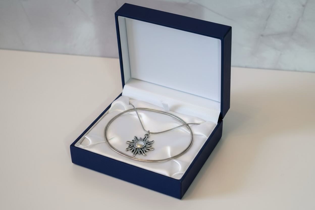 A silver-toned circular pendant necklace with a sunburst design and a central clear stone, displayed inside an open navy blue jewelry box with a white satin lining.