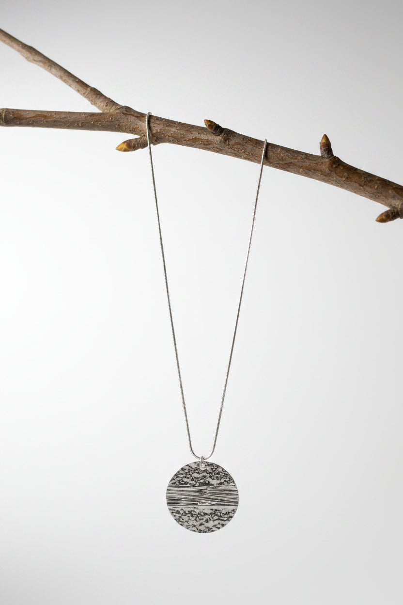 A silver pendant necklace with a textured circular medallion, displayed hanging over a tree branch.