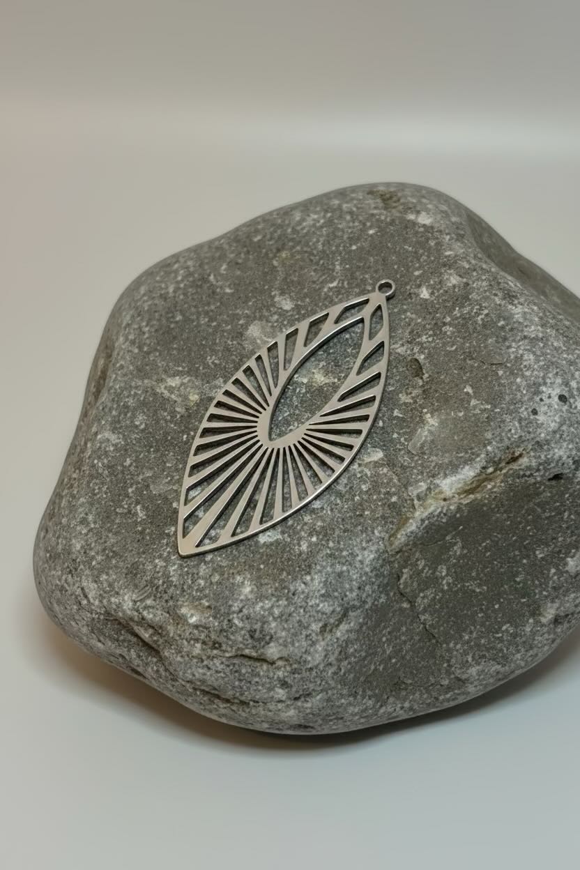 A silver-toned metal pendant with a leaf-like silhouette and radiating sunburst lines, displayed on a smooth gray stone against a light neutral background.