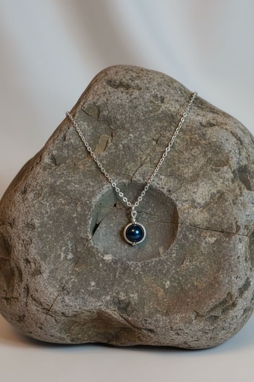 A delicate silver necklace with a steel blue round pearl pendant, resting on large grey stone against a light background.