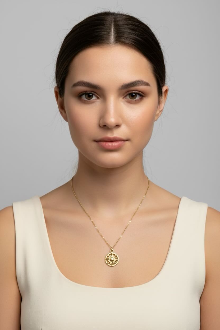 A woman with dark hair pulled back wears a delicate gold chain necklace with a circular sunburst pendant, paired with a cream-colored scoop-neck top, set against a solid gray studio background.