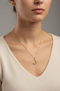 A close-up of a woman wearing a delicate gold crescent moon face pendant necklace with a small star detail, paired with a cream-colored knit top against a neutral gray background.