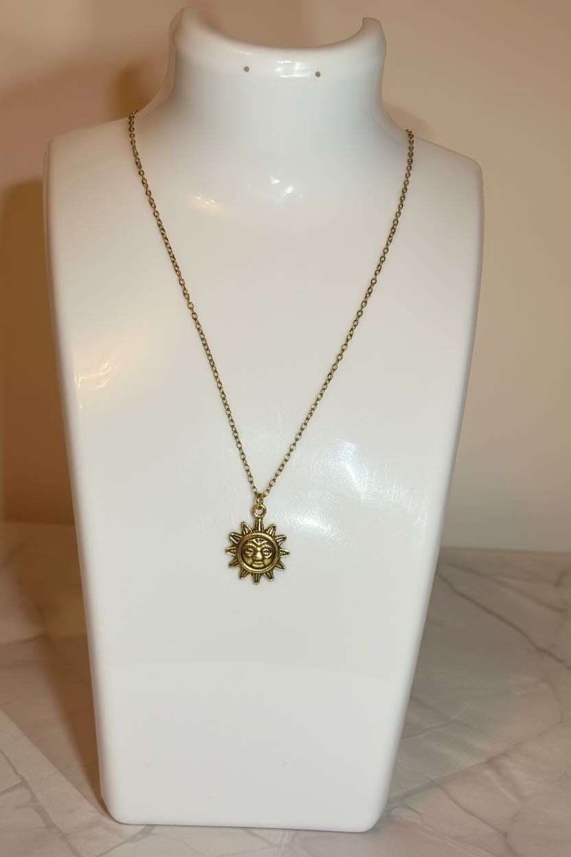 A gold-toned chain necklace with a sun face pendant featuring an anchor in the center, displayed on a white jewelry bust against a warm beige background.