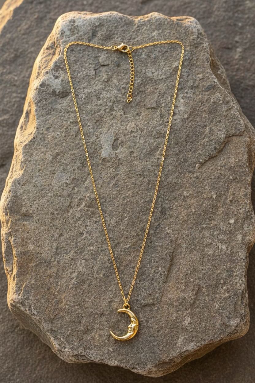 A delicate gold crescent moon face pendant necklace displayed on a textured gray stone surface.
