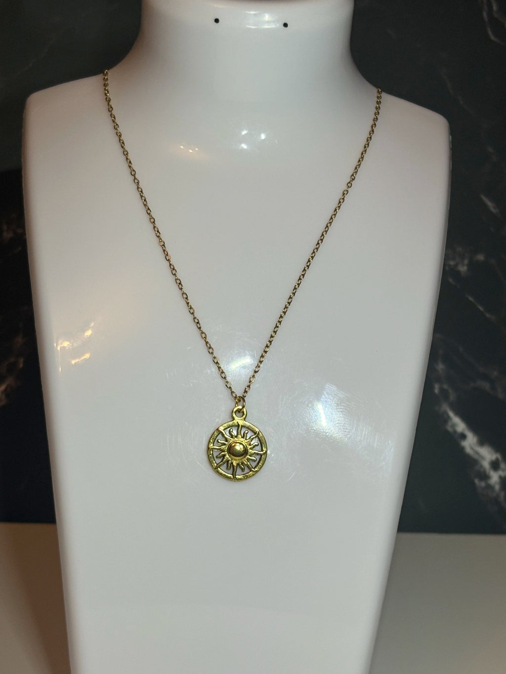 A gold-toned necklace with a circular sunburst pendant displayed on a white mannequin bust against a dark background.