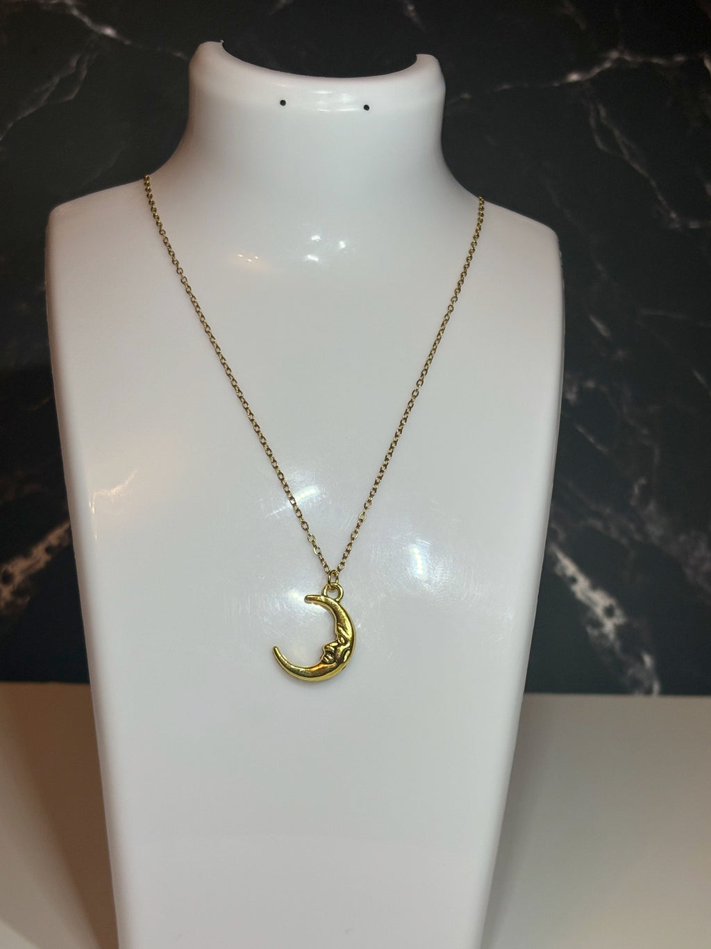 A gold crescent moon face pendant necklace on a delicate gold chain, displayed on a white mannequin bust against a dark marble-patterned background.