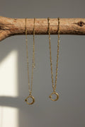 Two gold crescent moon necklaces with star details hanging from a rustic wooden branch against a neutral gray background.