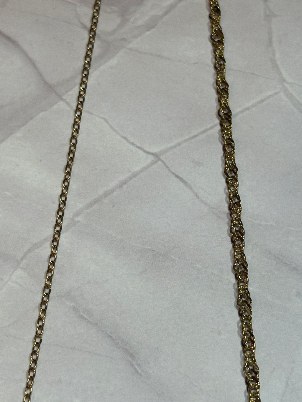 Two gold-toned metal chains laid flat on a wrinkled white surface, one with a fine cable-like link pattern and one with a slightly thicker curb-style link pattern.