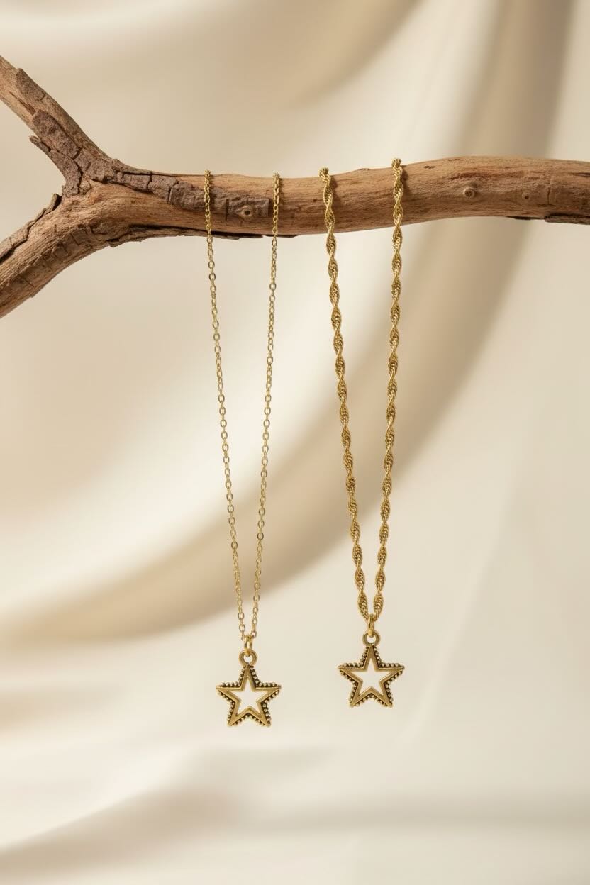 Two gold-toned star pendant necklaces hanging from a rustic wooden branch against a soft cream fabric backdrop.