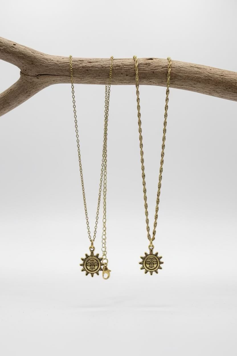 Two gold-toned chain necklaces with sun face pendants hanging from a natural wood branch against a light gray background.