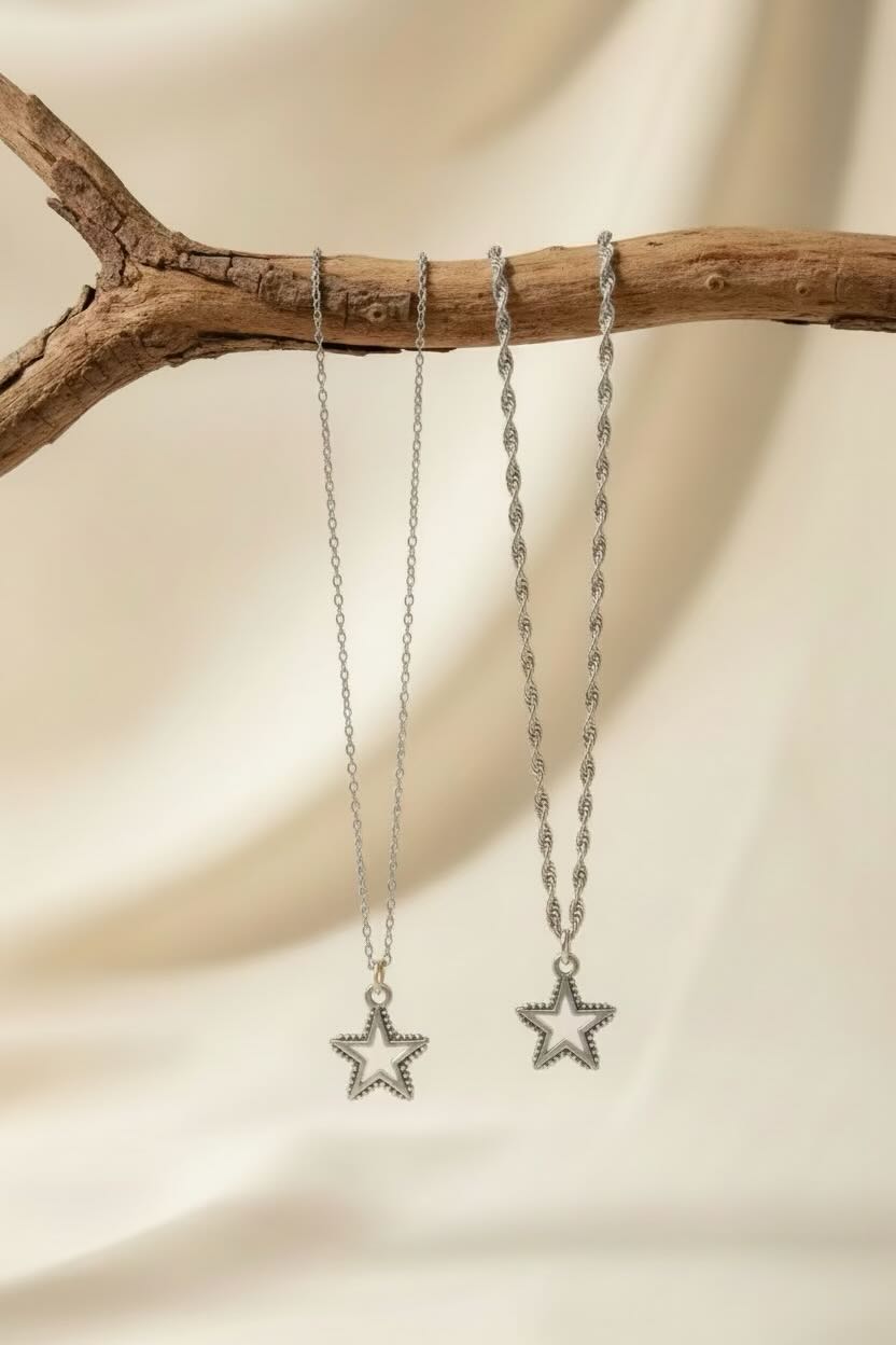 Two silver-toned star pendant necklaces hanging from a rustic wooden branch against a soft cream fabric backdrop.