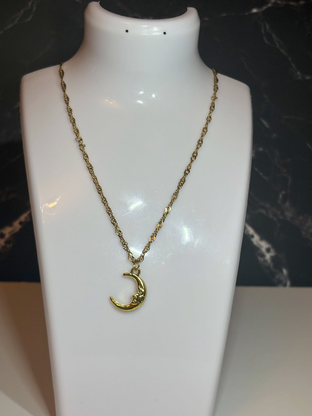A gold crescent moon face pendant necklace displayed on a white mannequin bust against a dark marbled background.