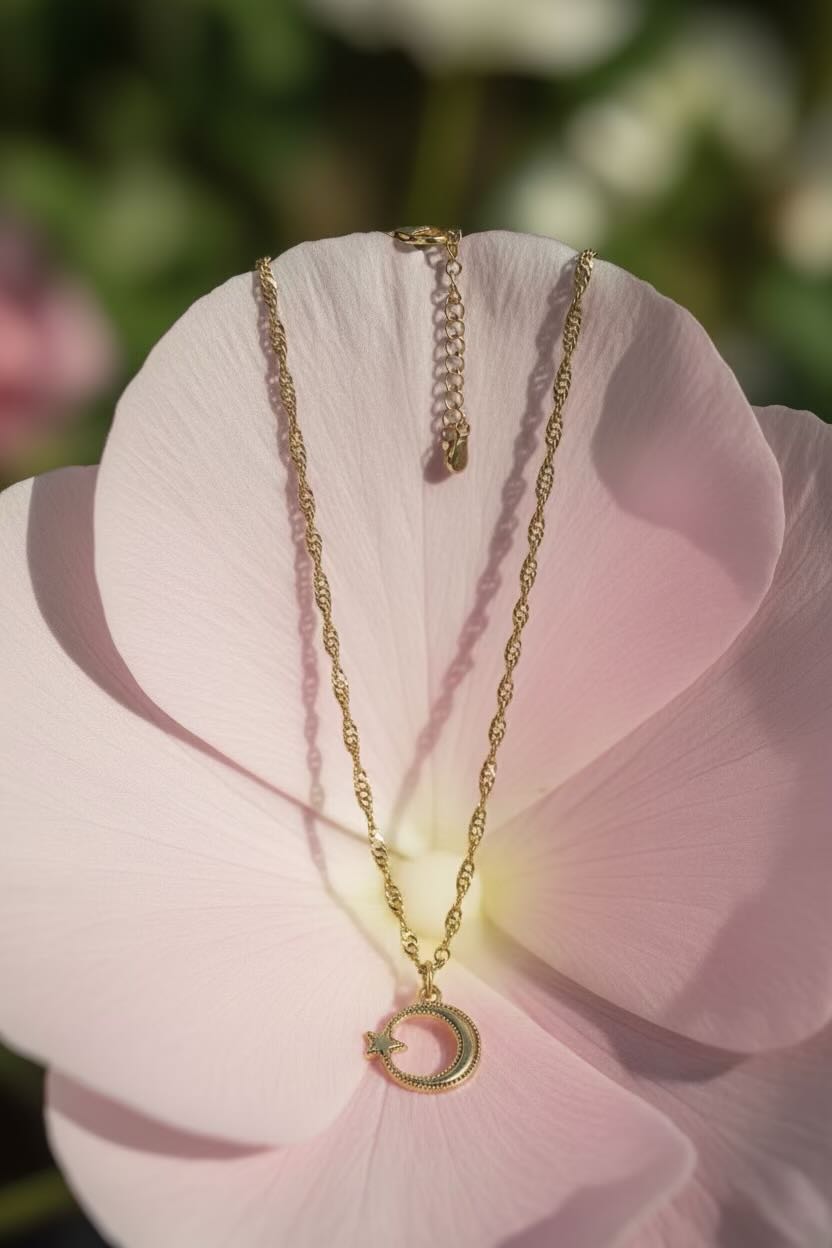 A delicate gold necklace with a crescent moon pendant rests on the center of a large light pink flower, with a soft, blurred green and white natural background.
